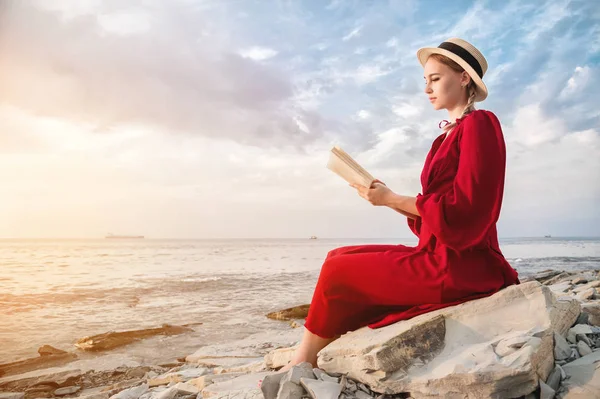 Atraktivní Kavkazská dívka v červených, světlých šatech a slaměném klobouku sedí na velkém kameni u moře při západu slunce a čte zajímavou knihu o mořském obzoru a nádherné mraky. — Stock fotografie