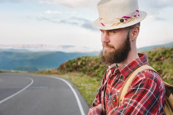 Retrato de um viajante sério feliz barbudo hipster com uma mochila em uma camisa xadrez e um chapéu ao lado de um carro desconhecido fica na estrada ao pôr do sol nas montanhas. Conceito de viagem feliz e confiante — Fotografia de Stock
