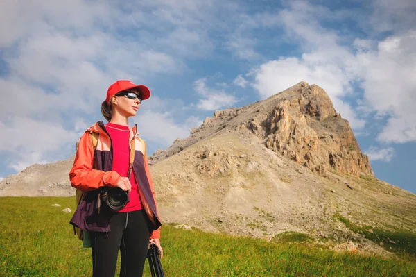 Menina fotógrafa atleta em um chapéu vermelho óculos de sol e uma mochila amarela fica em uma encosta verde contra o fundo das falésias épicas do Cáucaso — Fotografia de Stock
