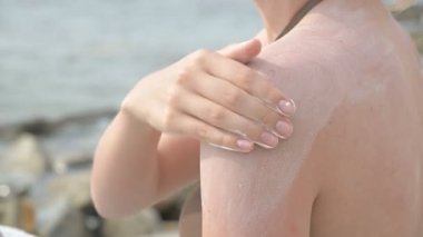 Güneşli bir yaz gününde deniz kıyısında otururken omzuna güneş kremi uygulayan çekici bir genç kız. Yaz koruma ve cilt bakım konsepti.