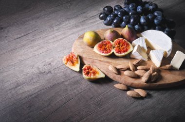 Ahşap bir kesme tahtası üzerinde Natürmort peynir plaka kabuk kalıp badem ve siyah üzüm bir demet yanında dilimlenmiş incir ile dilimlenmiş peynir yatıyor. Lezzetli ve sağlıklı gıda konsepti