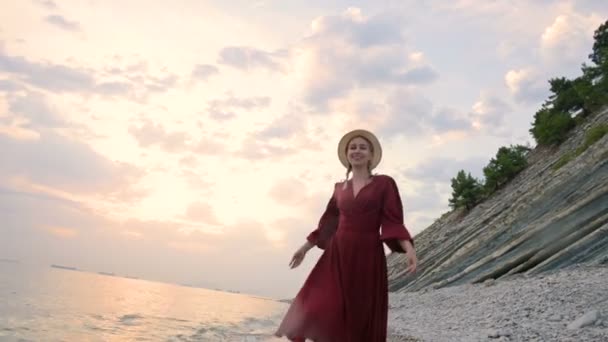 Joven chica sonriente atractiva en un vestido de luz roja y un sombrero de paja gira caminando a lo largo de la orilla del mar en tiempo ventoso al atardecer. Feminidad e independencia en ligereza y belleza — Vídeo de stock