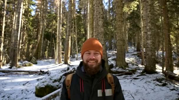 Ritratto di un giovane allegro barbuto che ride con uno zaino in piedi in una foresta invernale di conifere — Video Stock
