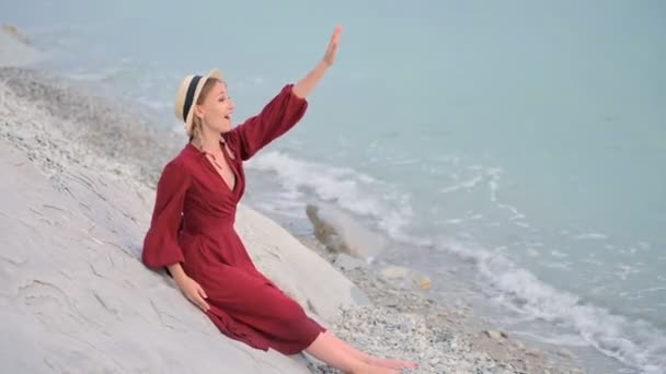 Una chica sonriente feliz con un vestido rojo de verano y un sombrero de paja se sienta en una orilla rocosa y mira el horizonte del mar. Agita sus manos, llamando la atención sobre sí mismo . — Vídeos de Stock