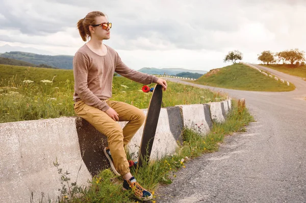 Homem elegante jovem com cabelos longos em óculos de sol está sentado em um chipper com um longboard em suas mãos em uma estrada de asfalto país no fundo de rochas e nuvens — Fotografia de Stock