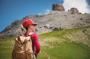 Kırmızı şapkalı güneş gözlüğü ve sarı sırt çantası ile arka Kız atlet görünümü Kafkasya'nın epik kayalıkların arka plan karşı yeşil bir yamaç üzerinde duruyor