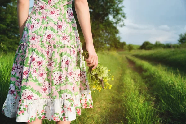 Uma vista da parte de trás de uma menina em um vestido de verão leve com um buquê de flores silvestres está em frente a uma estrada rural. O conceito de feminismo de igualdade de direitos e independência das mulheres — Fotografia de Stock