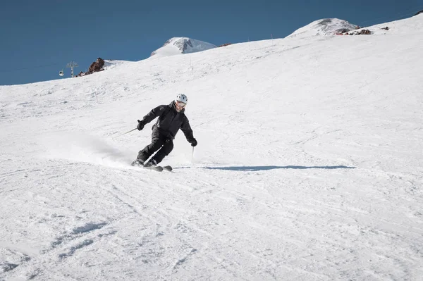 A bearded mature aged male skier in a black ski suit descends along the snowy slope of a ski resort amid two peaks of Mount Elbrus. The concept of sports in adulthood
