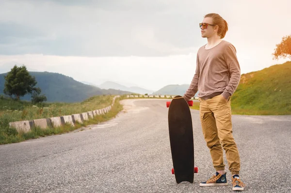 Jovem elegante de pé ao longo de uma estrada de montanha sinuosa com um skate ou longboard em seu ombro à noite após o pôr do sol. O conceito de esportes juvenis e hobbies de viagem — Fotografia de Stock