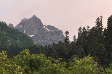 Bulutlar tepeye sarılmış karanlık bulutlu gökyüzüne karşı kısmen karla kaplı yüksek bir dik kayalık dağ görünümü. Dağ ormanı hava değişikliği kavramı
