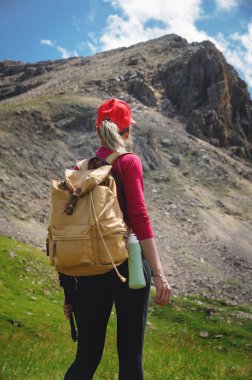 Kırmızı şapkalı güneş gözlüğü ve sarı sırt çantası ile arka Kız atlet görünümü Kafkasya'nın epik kayalıkların arka plan karşı yeşil bir yamaç üzerinde duruyor