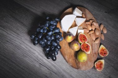 Ahşap bir kesme tahtası üzerinde Natürmort peynir plaka kabuk kalıp badem ve siyah üzüm bir demet yanında dilimlenmiş incir ile dilimlenmiş peynir yatıyor. Lezzetli ve sağlıklı gıda konsepti