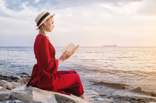 Ένα ελκυστικό καυκάσιο κορίτσι σε ένα κόκκινο φωτεινό φόρεμα και ένα ψάθινο καπέλο κάθεται σε μια μεγάλη πέτρα δίπλα στη θάλασσα το ηλιοβασίλεμα και διαβάζει ένα ενδιαφέρον βιβλίο στον ορίζοντα της θάλασσας και όμορφα σύννεφα. — Φωτογραφία Αρχείου