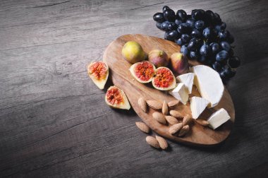 Ahşap bir kesme tahtası üzerinde Natürmort peynir plaka kabuk kalıp badem ve siyah üzüm bir demet yanında dilimlenmiş incir ile dilimlenmiş peynir yatıyor. Lezzetli ve sağlıklı gıda konsepti