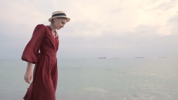 Молодая привлекательная улыбающаяся девушка в красном светлом платье и соломенной шляпе прогуливается по берегу моря в ветреную погоду на закате. Женственность и независимость в легкости и красоте — стоковое видео