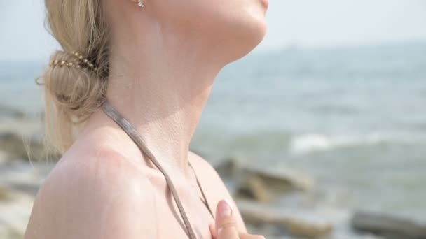 Primer plano de tiro. Una joven atractiva con gafas de sol que se aplica protector solar en el cuello mientras está sentada en la orilla del mar en un día soleado de verano. Protección de verano y concepto de cuidado de la piel . — Vídeo de stock