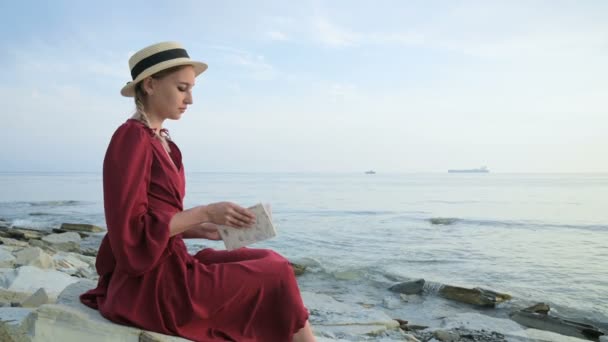 Uma menina caucasiana atraente em um vestido vermelho brilhante e um chapéu de palha senta-se em uma grande pedra junto ao mar ao pôr do sol e lê um livro interessante no horizonte do mar e belas nuvens . — Vídeo de Stock