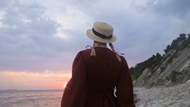 La vista desde atrás. Retrato en cámara lenta de una joven con un vestido rojo y un sombrero de paja en la orilla del mar. Una chica en el viento sostiene un sombrero con sus manos. Esperando el regreso de los marineros — Vídeos de Stock