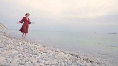 Kırmızı ışık elbise ve hasır şapka genç bir çekici gülümseyen kız gün batımında rüzgarlı havalarda deniz kıyısında yürür. Hafiflik ve güzellikte kadınlık ve bağımsızlık