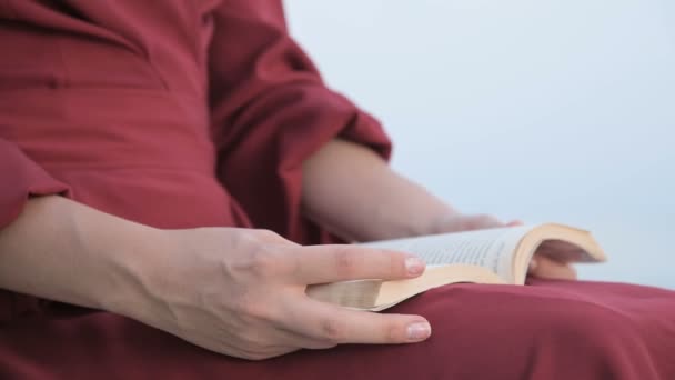 Close-up uma menina caucasiana atraente mãos em um vestido vermelho brilhante e um chapéu de palha senta-se em uma grande pedra junto ao mar ao pôr do sol e lê um livro interessante — Vídeo de Stock