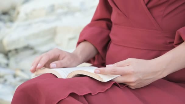 Close-up uma menina caucasiana atraente mãos em um vestido vermelho brilhante e um chapéu de palha senta-se em uma grande pedra junto ao mar ao pôr do sol e lê um livro interessante — Vídeo de Stock