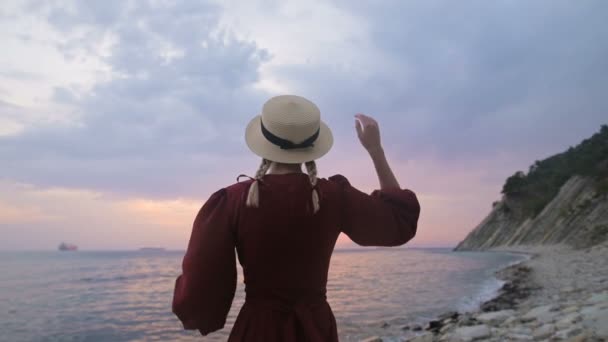 後ろから見た眺め。赤いドレスと海岸に麦わら帽子をかぶった少女のスローモーションポートレート。風の中の少女は手で帽子を持っている。船員の帰還を待っている — ストック動画