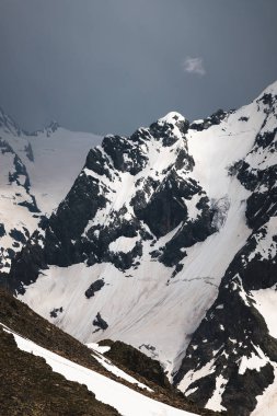 Bulutlar tepeye sarılmış karanlık bulutlu gökyüzüne karşı kısmen karla kaplı yüksek bir dik kayalık dağ görünümü. Dağ hava sıcamı kavramı