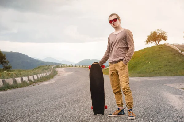 Um jovem elegante de pé ao longo de uma estrada de montanha sinuosa com um skate ou longboard em suas mãos na noite após o pôr do sol. O conceito de esportes juvenis e hobbies de viagem — Fotografia de Stock