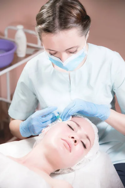 Jovem atraente recebe injeções faciais anti-envelhecimento. Ela se deita calmamente em uma clínica ou salão. Um jovem cosmetologista experiente enche rugas femininas com ácido hialurônico de uma seringa — Fotografia de Stock