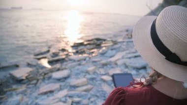 Yakın plan görüntüsü, yaz aylarında kırmızı elbiseli ve hasır şapkalı çekici bir kız gün batımında deniz kenarında bir taşın üzerinde oturup tablet üzerinde bir şey izliyor. Ekran boyunca kaydır