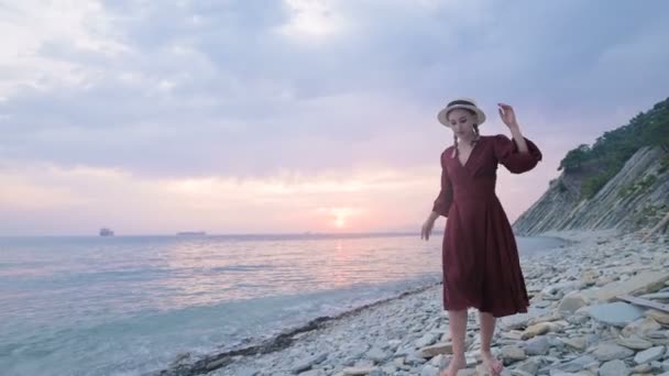 Une jolie jeune fille en robe d'été rouge et un chapeau de paille glisse prudemment sur les pierres du rivage rocheux de la mer sur fond de vagues et de coucher de soleil . — Video