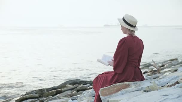 Vista posterior Una atractiva chica caucásica en un vestido rojo brillante y un sombrero de paja se sienta en una gran piedra junto al mar al atardecer y lee un interesante libro sobre el horizonte del mar y las hermosas nubes . — Vídeos de Stock