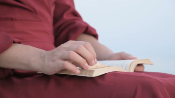 Gros plan une belle fille caucasienne mains dans une robe rouge vif et un chapeau de paille se trouve sur une grande pierre au bord de la mer au coucher du soleil et lit un livre intéressant — Video