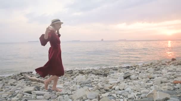 Una joven atractiva con un vestido rojo de verano y un sombrero de paja se arrastra cautelosamente sobre las piedras de la costa rocosa del mar contra el fondo de las olas y la puesta de sol . — Vídeos de Stock