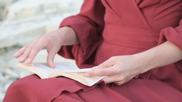 Close-up uma menina caucasiana atraente mãos em um vestido vermelho brilhante e um chapéu de palha senta-se em uma grande pedra junto ao mar ao pôr do sol e lê um livro interessante — Vídeo de Stock