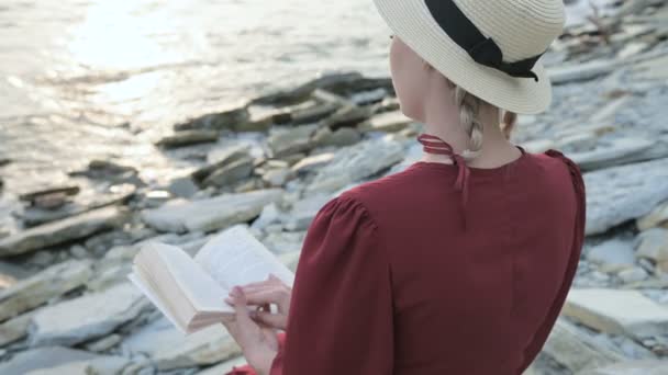 Vista trasera Close-up Una atractiva chica caucásica en un vestido rojo brillante y un sombrero de paja se sienta en una gran piedra junto al mar al atardecer y lee un libro interesante — Vídeos de Stock