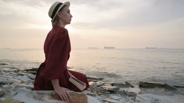 Una atractiva chica caucásica en un vestido rojo brillante y un sombrero de paja se sienta en una gran piedra junto al mar al atardecer y lee un interesante libro sobre el horizonte del mar y las hermosas nubes . — Vídeos de Stock