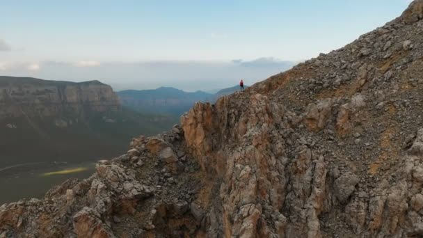 Vista aérea de uma menina viajante com uma mochila fica à beira de um penhasco de uma alta montanha rochosa e olha para as montanhas cobertas de neve à distância. Viagens vídeo drone footage — Vídeo de Stock