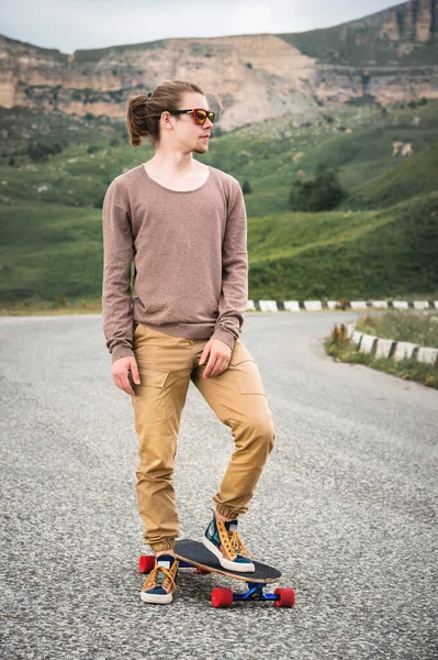 Um jovem elegante de pé ao longo de uma estrada de montanha sinuosa com um skate ou longboard em suas mãos na noite após o pôr do sol. O conceito de esportes juvenis e hobbies de viagem — Fotografia de Stock