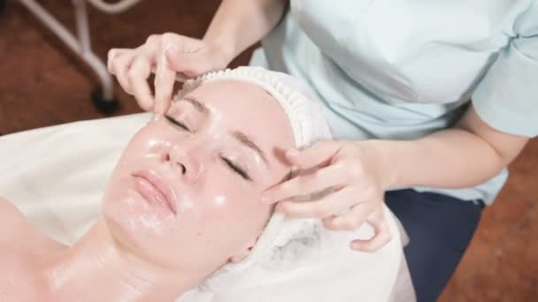 Menina esteticista em uma máscara e luvas faz uma massagem facial salubre para uma mulher atraente. Nova geração massagem cosmetologia — Vídeo de Stock