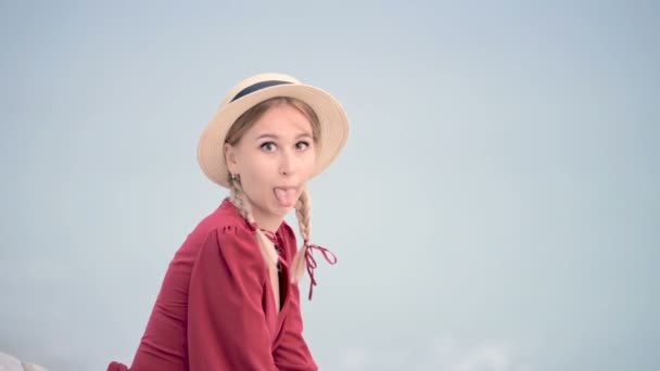 Portrait attractive caucasian young woman in a red dress and a straw hat with pigtails sitting on the seashore smiling looking at the camera and grimacing while making fun showing tongue — Stock Video