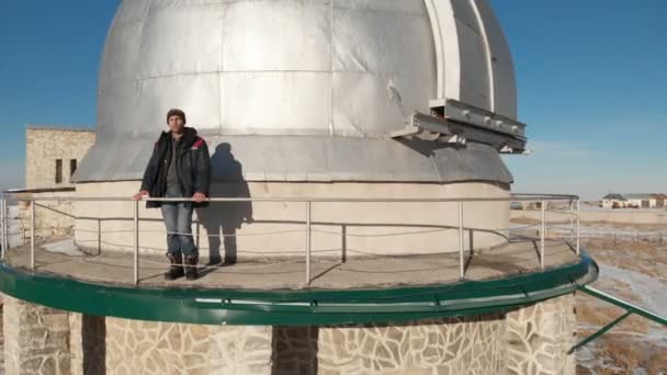 An adult male observatory worker stands on the observation deck of the observation dome and looks into the distance. Aerial view against a clear blue sky. Astronomy science concept — ストック動画