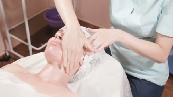 Chica esteticista en una máscara y guantes hace un masaje facial saludable a una mujer atractiva. Masaje cosmetológico de nueva generación — Vídeos de Stock