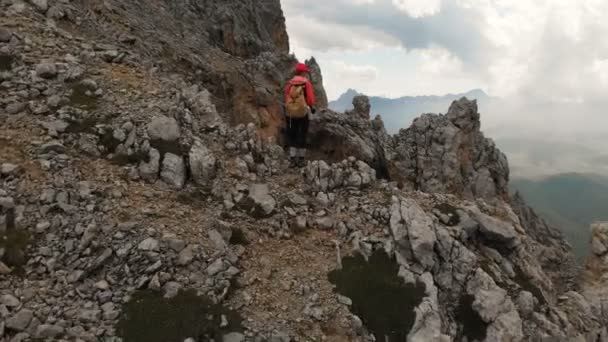 Letecký pohled na dívku cestovatele s batohem stojí na okraji útesu vysoké skalnaté hory a dívá se na zasněžené hory v dálce. Cestovní video drone záběry — Stock video
