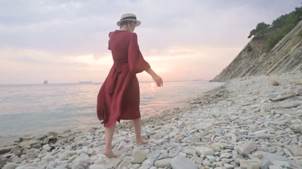 An attractive young girl in a red summer dress and a straw hat cautiously creeps on the stones of the sea rocky shore against the background of waves and sunset. — Stock Video