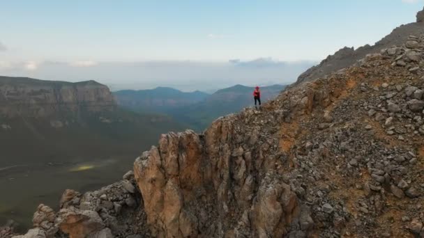 Luchtfoto van een meisje reiziger met een rugzak staat op de rand van een klif van een hoge rotsachtige berg en kijkt naar de besneeuwde bergen in de verte. Reisvideo drone beelden — Stockvideo