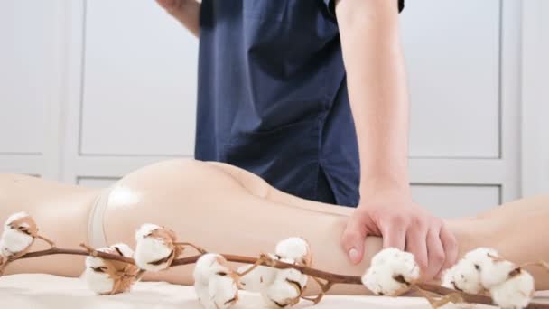 Nahaufnahme eines männlichen Physiotherapeuten, der einer jungen Frau vor Massageanwendungen in einem professionellen Wellness-Salon Massageöl auf den Oberschenkel und das Gesäß reibt. Konzept der Entspannung und Körperpflege in 4k — Stockvideo