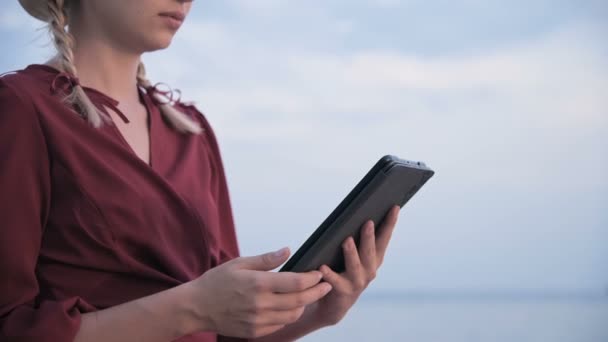 Close-up An attractive young girl hands in a summer red dress sits on a stone by the sea in the evening and looks at something on a tablet. Swipe across the screen — ストック動画