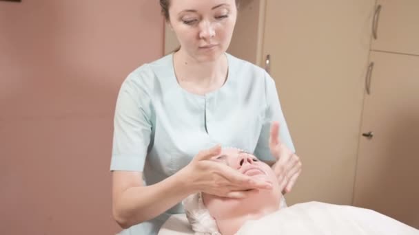 Дівчина-косметолог у масці та рукавичках робить солоний масаж обличчя привабливою жінкою. Косметологічний масаж нового покоління — стокове відео