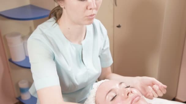 Menina esteticista em uma máscara e luvas faz uma massagem facial salubre para uma mulher atraente. Nova geração massagem cosmetologia — Vídeo de Stock